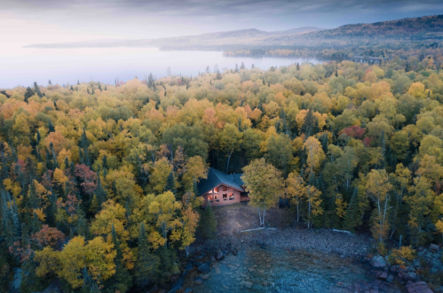 Aerial view of a cozy cabin surrounded by vibrant autumn foliage near a serene lake.