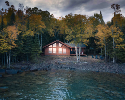 A cozy cabin by a lake, surrounded by autumn trees and rocky shoreline under a cloudy sky.