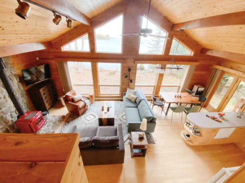 Cozy cabin interior with wooden beams, large windows, a stone fireplace, and a mix of seating and dining areas.