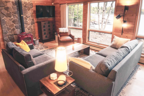 Cozy living room with wooden walls, large windows, a stone fireplace, and comfortable seating overlooking a lake.