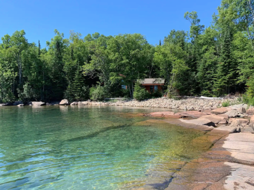 A serene lakeside scene with clear water, rocky shore, and a cabin nestled among lush green trees under a blue sky.