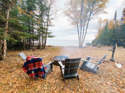 A cozy campsite with chairs around a fire pit, surrounded by autumn leaves and trees near a lake.