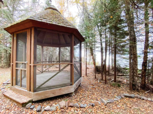 A wooden gazebo surrounded by trees, with a stone path and fallen leaves on the ground.