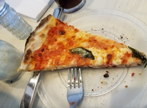 A slice of pizza with cheese, tomato sauce, and basil on a clear plate, accompanied by a fork and knife.