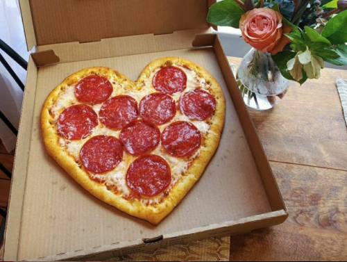 A heart-shaped pepperoni pizza in a pizza box, next to a vase of flowers on a wooden table.