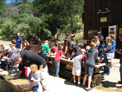 A group of children and adults are panning for gold at a historic site, surrounded by trees and a rustic building.