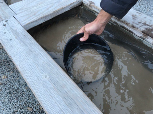 A hand holds a black bucket submerged in muddy water, next to a wooden structure.