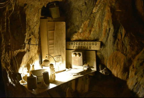 A dimly lit underground workspace with a wooden table, old tools, and a sign reading "Underground Foreman."