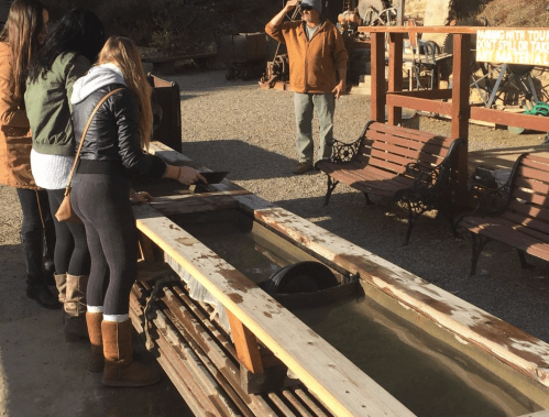 A group of people panning for gold at a wooden trough, with a guide observing in the background.