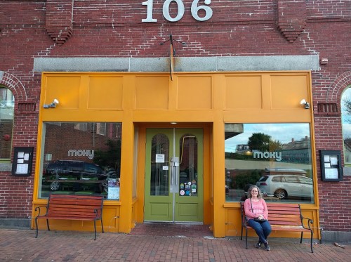 A woman sits on a bench outside a bright orange storefront with large windows and the name "Moky" displayed.