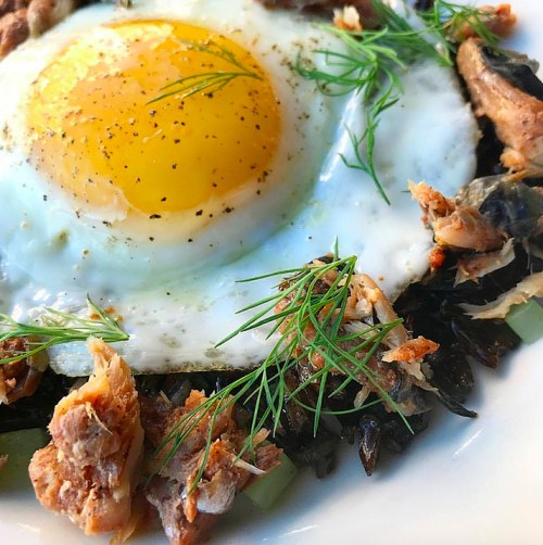 A close-up of a dish featuring a fried egg on a bed of black rice, topped with herbs and pieces of fish.