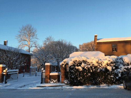 A snowy scene featuring a house and trees, with sunlight illuminating the snow-covered landscape.