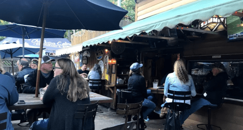 A lively outdoor bar scene with patrons sitting at tables and a bar, surrounded by umbrellas and festive lights.