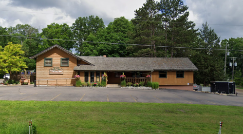 A wooden building with a sloped roof, surrounded by trees and greenery, featuring flower pots and a parking area.