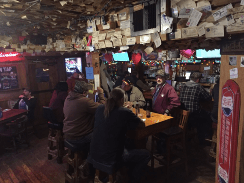 A cozy bar scene with patrons sitting at wooden tables, surrounded by colorful lights and decorated walls.