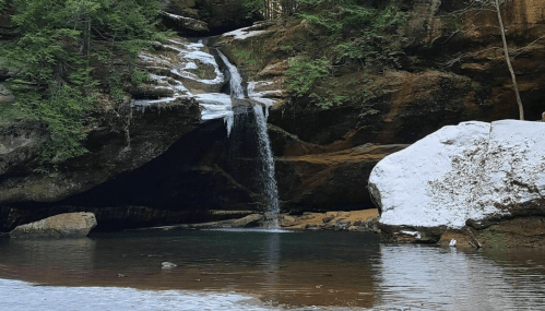 A serene waterfall cascades over rocks into a calm pool, surrounded by snow and lush greenery.