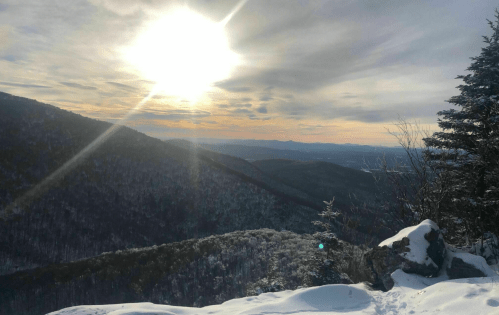 A snowy mountain landscape under a bright sun, with distant hills and a cloudy sky in the background.