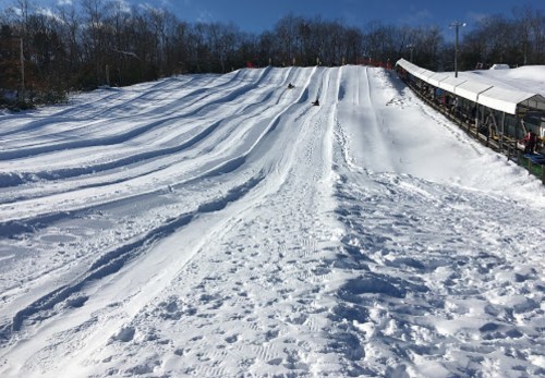 A snowy hill with sledding tracks and a covered seating area in the background under a clear blue sky.