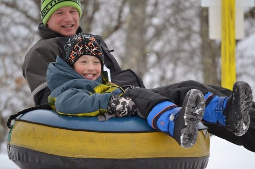 A man and a boy smile while sitting on a snow tube, enjoying a winter day in a snowy landscape.