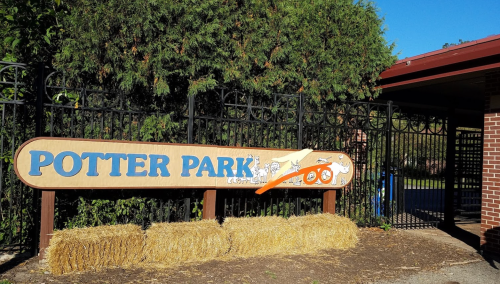 Sign for Potter Park Zoo with a wooden design, surrounded by greenery and a fence. Hay bales are in front of the sign.
