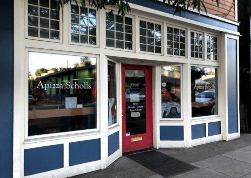 Exterior of Apizza Scholls, featuring large windows and a red door, with "OPEN" sign and a welcoming atmosphere.