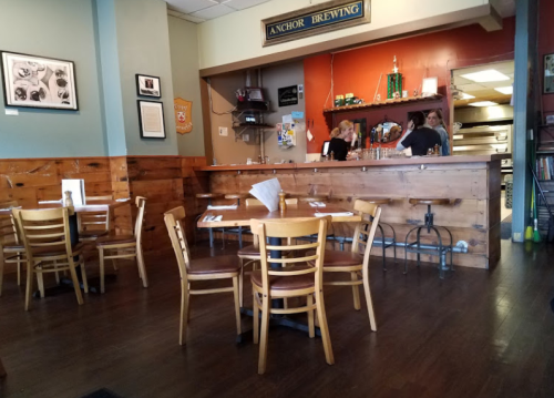 Interior of a casual restaurant with wooden tables, a bar area, and two staff members behind the counter.
