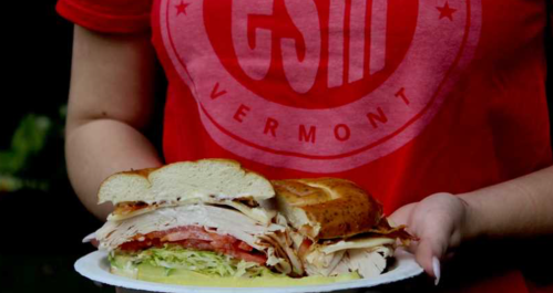 A person holding a large sandwich with turkey, lettuce, and tomato on a plate, wearing a red shirt with "Vermont" printed on it.