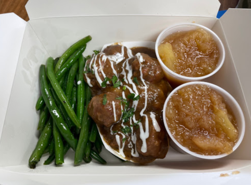 A takeout box containing green beans, meatballs with sauce, and two cups of applesauce.