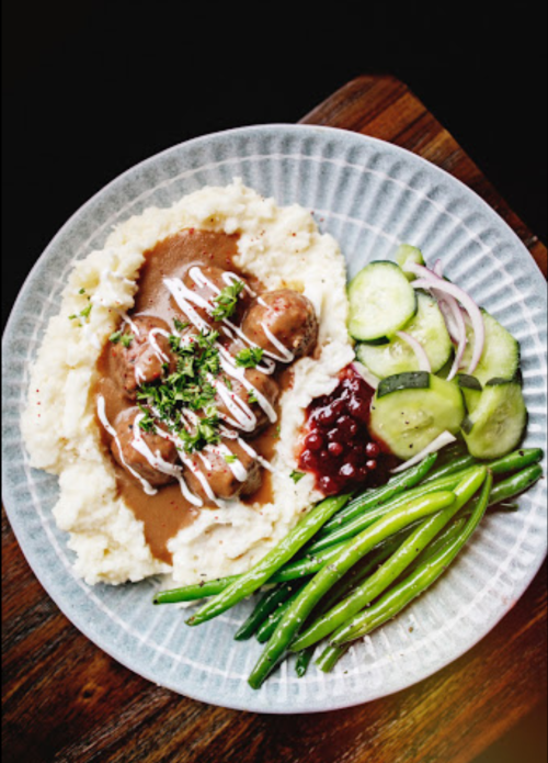 A plate of mashed potatoes with gravy, green beans, pickled cucumbers, and a side of red sauce.