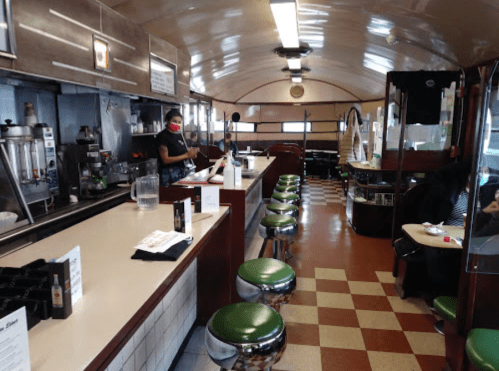 A retro diner interior with green stools, a counter, and a staff member wearing a mask. Checkered floor and booths visible.
