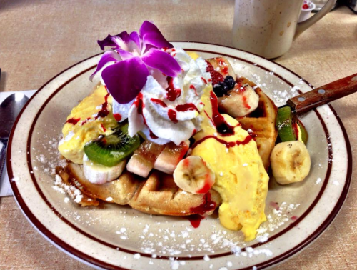 A plate of waffles topped with bananas, kiwi, whipped cream, syrup, and a purple flower garnish.