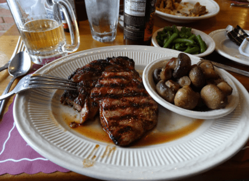 Grilled steak with a side of mushrooms and green beans, accompanied by a glass of beer and a water glass.