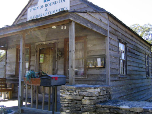 A rustic wooden building with a sign for the Town of Round Top and Chamber of Commerce, featuring a stone wall in front.