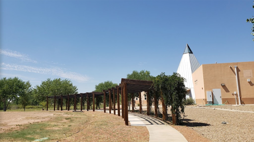 A pathway lined with wooden trellises leads to a building with a conical roof under a clear blue sky.