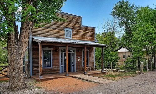 A rustic wooden building with a porch, surrounded by trees and a gravel road under a clear blue sky.