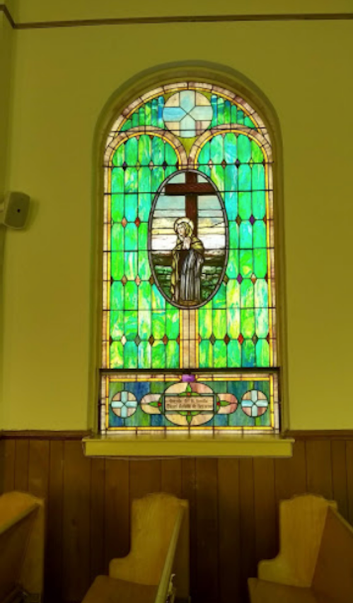 Stained glass window featuring a figure in front of a cross, surrounded by vibrant green and blue patterns.