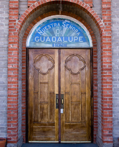 Wooden double doors with an arched stained glass window above, featuring "Nuestra Señora de Guadalupe 1996" in blue.