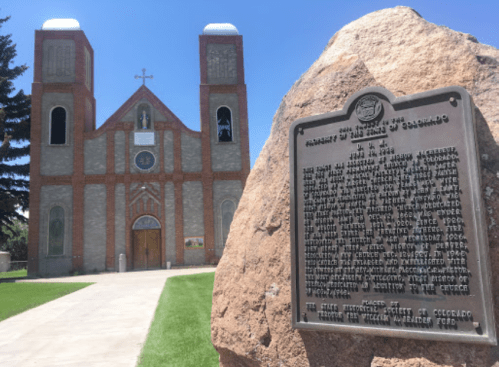 Historic church with a stone plaque in front, surrounded by green grass and trees under a clear blue sky.