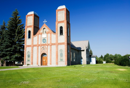 A brick church with two towers, surrounded by green grass and trees, under a clear blue sky.