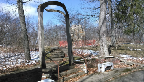 An old, weathered archway stands at the top of stone steps in a wooded area, with remnants of snow and a distant building.