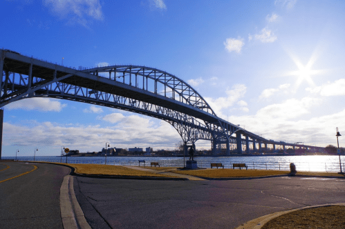 A large steel bridge spans over a river, with a clear blue sky and sunlight shining in the background. Benches line the shore.