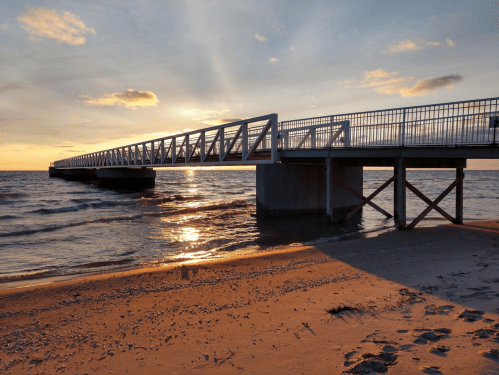 A wooden pier extends over calm waters at sunset, with soft waves lapping against the sandy shore.