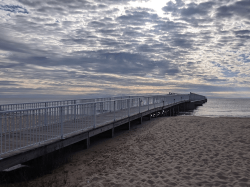 A wooden pier extends into a calm sea under a cloudy sky, with sandy beach in the foreground.