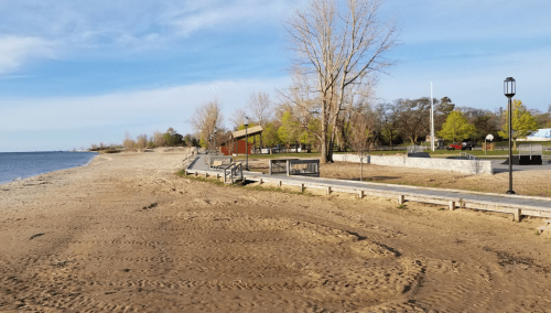 A sandy beach with a wooden walkway, trees, and a building in the background under a clear blue sky.