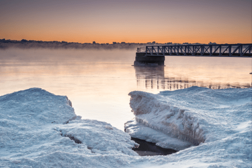 A serene winter scene with icy formations by a calm river at sunrise, featuring a pier in the background.
