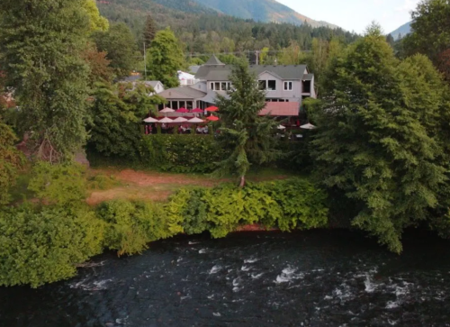 Aerial view of a riverside restaurant surrounded by lush greenery and mountains in the background.