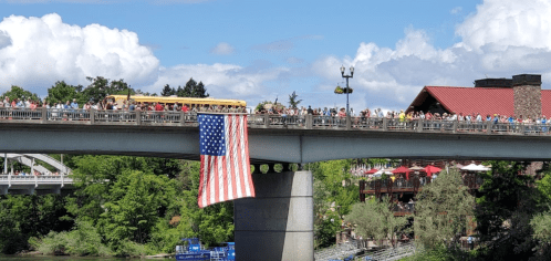 A large crowd gathers on a bridge, with an American flag hanging down and a scenic backdrop of trees and buildings.
