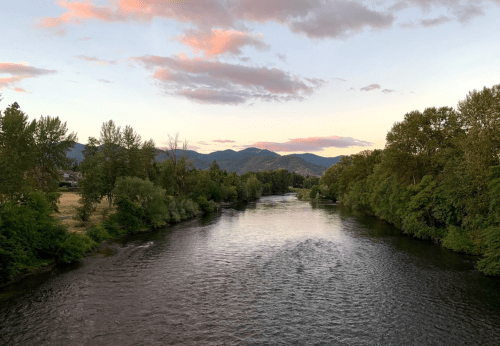 A serene river flows through lush greenery, with mountains and a colorful sunset in the background.