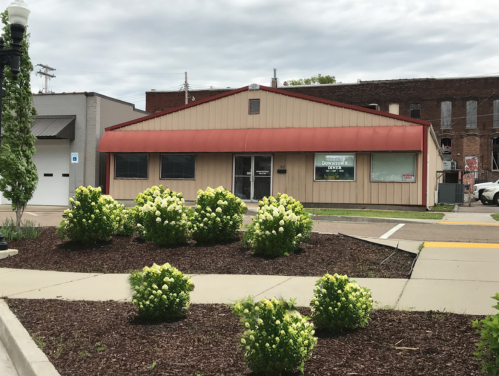 A single-story building with a red roof, surrounded by green shrubs and flowers, under a cloudy sky.