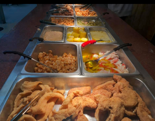 A buffet spread featuring various dishes, including fried seafood, corn, and sauces in metal trays with serving utensils.